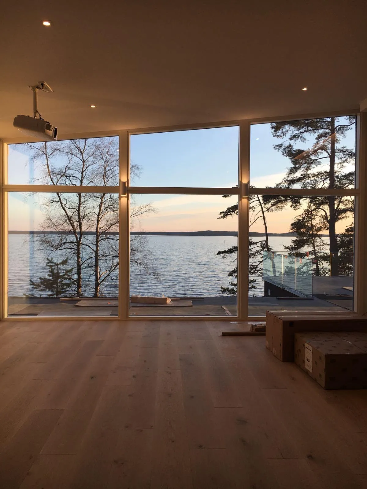 Sisäkuva huoneesta, jossa on suuret ikkunat järvelle päin auringonlaskun aikaan. Lattialla on vaalea puulattia ja katossa upotettuja valoja sekä videoprojektori. Ikkunoista näkyy tyyni järvi, paljaita puita vasemmalla ja mäntyjä oikealla. Taivas on vaaleansininen, oranssi ja vaaleanpunainen. Oikealla alhaalla on pahvilaatikoita ja rakennustarvikkeita.