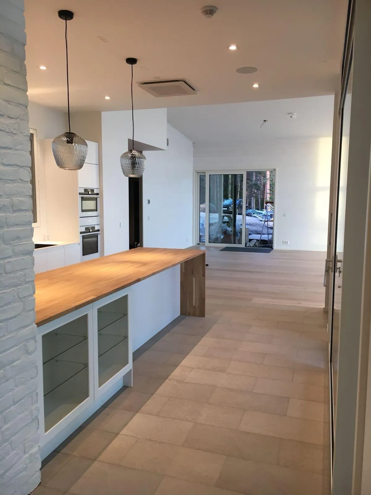 Moderni avokeittiö ja olohuone, jossa on valkoiset seinät ja vaalea puulattia. Keittiösaarekkeessa on vaalea puinen työtaso ja valkoiset kaapit lasiovilla. Kaksi hopeanväristä, kuvioitua lasista riippuvalaisinta roikkuu saarekkeen yläpuolella. Katossa on upotettuja kohdevaloja ja ilmastointikanava. Suuret lasiset liukuovet avautuvat ulos metsäiseen maisemaan. Lattialla on vaaleanruskeita laattoja keittiön alueella ja vaaleita puulankkuja olohuoneessa.