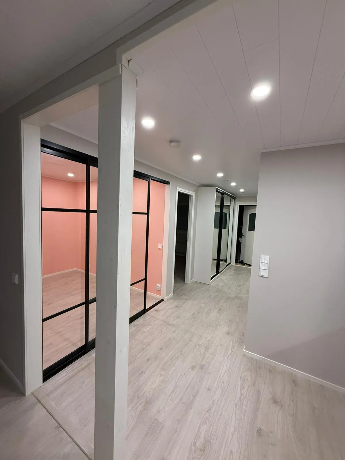 Sisäkuva käytävästä, jossa on vaalea puunvärinen laminaattilattia ja valkoinen paneelikatto upotetuilla kohdevaloilla. Vasemmalla näkyy vaaleanpunainen huone mustakehyksisten lasiovien takana. Edessä on valkoinen pylväs. Oikealla on vaaleanharmaa seinä valkoisella valokatkaisimella. Käytävän perällä on valkoinen vaatekaappi peiliovilla ja toinen valkoinen ovi. Katossa on myös palovaroitin. Tila on kirkkaasti valaistu.