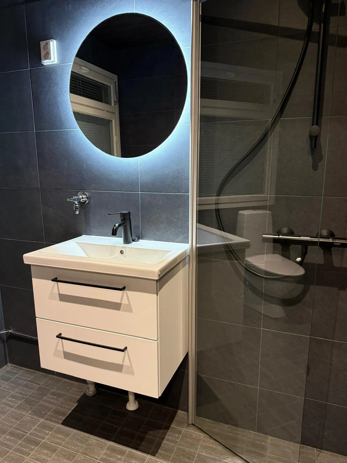 Moderni kylpyhuone, jossa on valkoinen allaskaappi pesualtaalla ja mustalla hanalla. Kaapin yläpuolella on pyöreä, taustavalaistu peili ja vasemmalla seinällä valaistu pistorasia. Tummanharmaat laatat peittävät seinät ja lattian. Oikealla näkyy lasinen suihkuseinä, joka heijastaa WC-istuimen ja suihkuvarusteet.