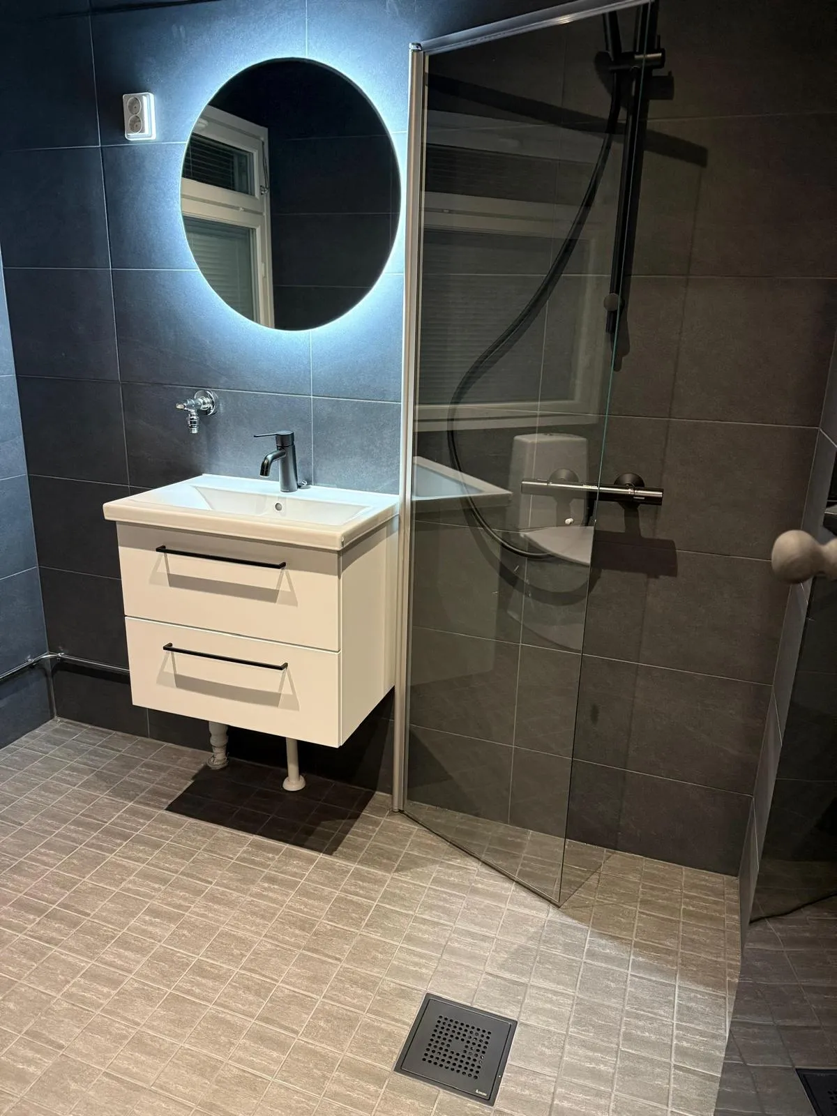 Moderni kylpyhuone tummanharmailla seinälaatoilla ja vaaleanharmailla lattialaatoilla. Vasemmalla on pyöreä, taustavalaistu peili, jonka alla on valkoinen allaskaappi pesualtaalla ja mustalla hanalla. Kaapissa on kaksi laatikkoa mustilla vetimillä. Oikealla on lasinen suihkuseinä, jonka takana näkyy musta suihku ja harmaa hylly. Lattialla on tummanharmaa lattiakaivo.