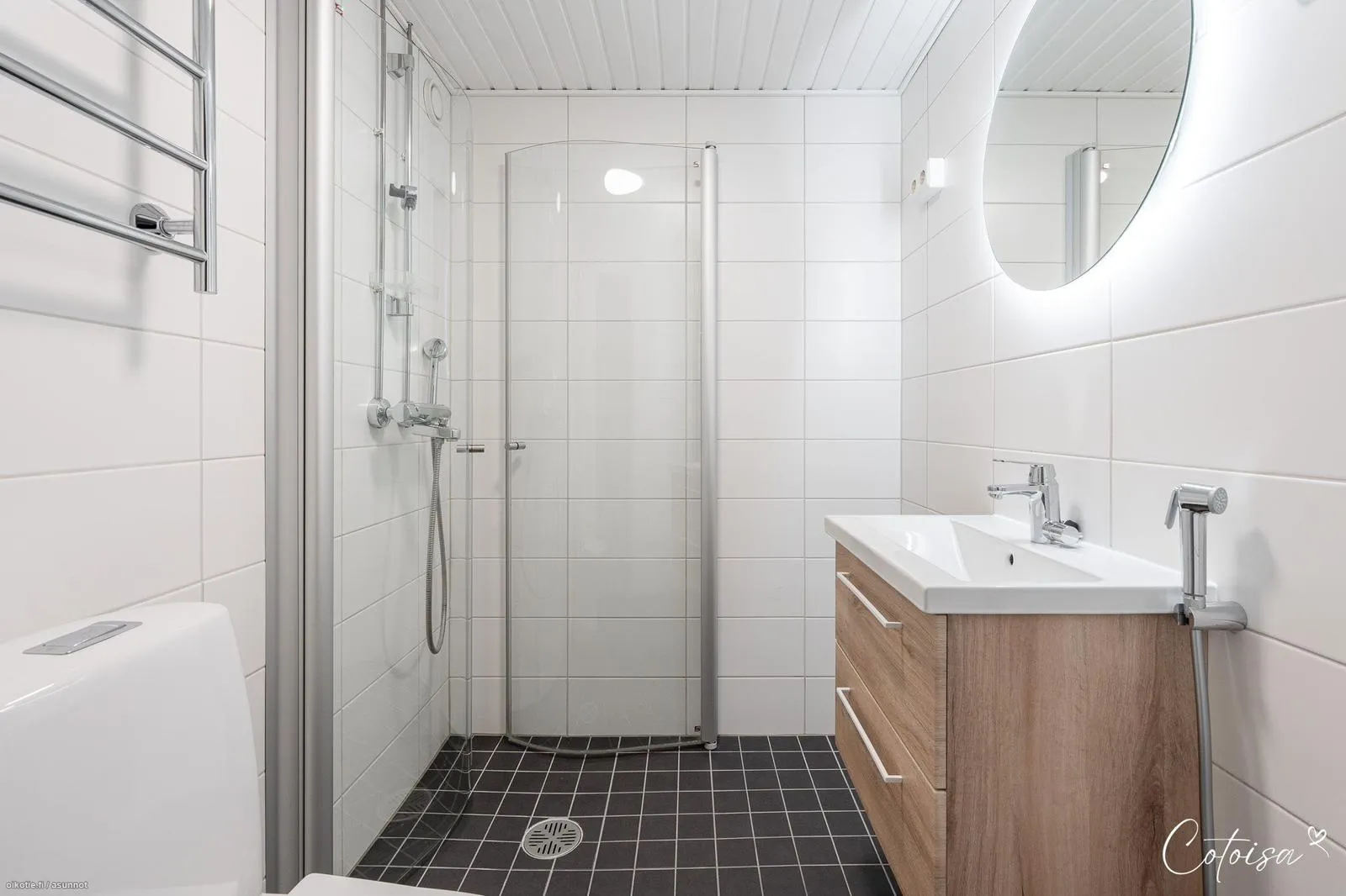 Moderni kylpyhuone, jossa on valkoiset laattaseinät ja tummanharmaat laattalattiat. Vasemmalla näkyy valkoinen WC-istuin ja krominen pyyhekuivain. Keskellä on lasiseinäinen suihkutila kromisella suihkulla. Oikealla on vaaleapuinen allaskaappi valkoisella altaalla, kromisella hanalla ja bideesuihkulla. Allaskaapin yläpuolella on pyöreä, valaistu peili. Katossa on valkoinen rimakatto ja seinällä pistorasiat.