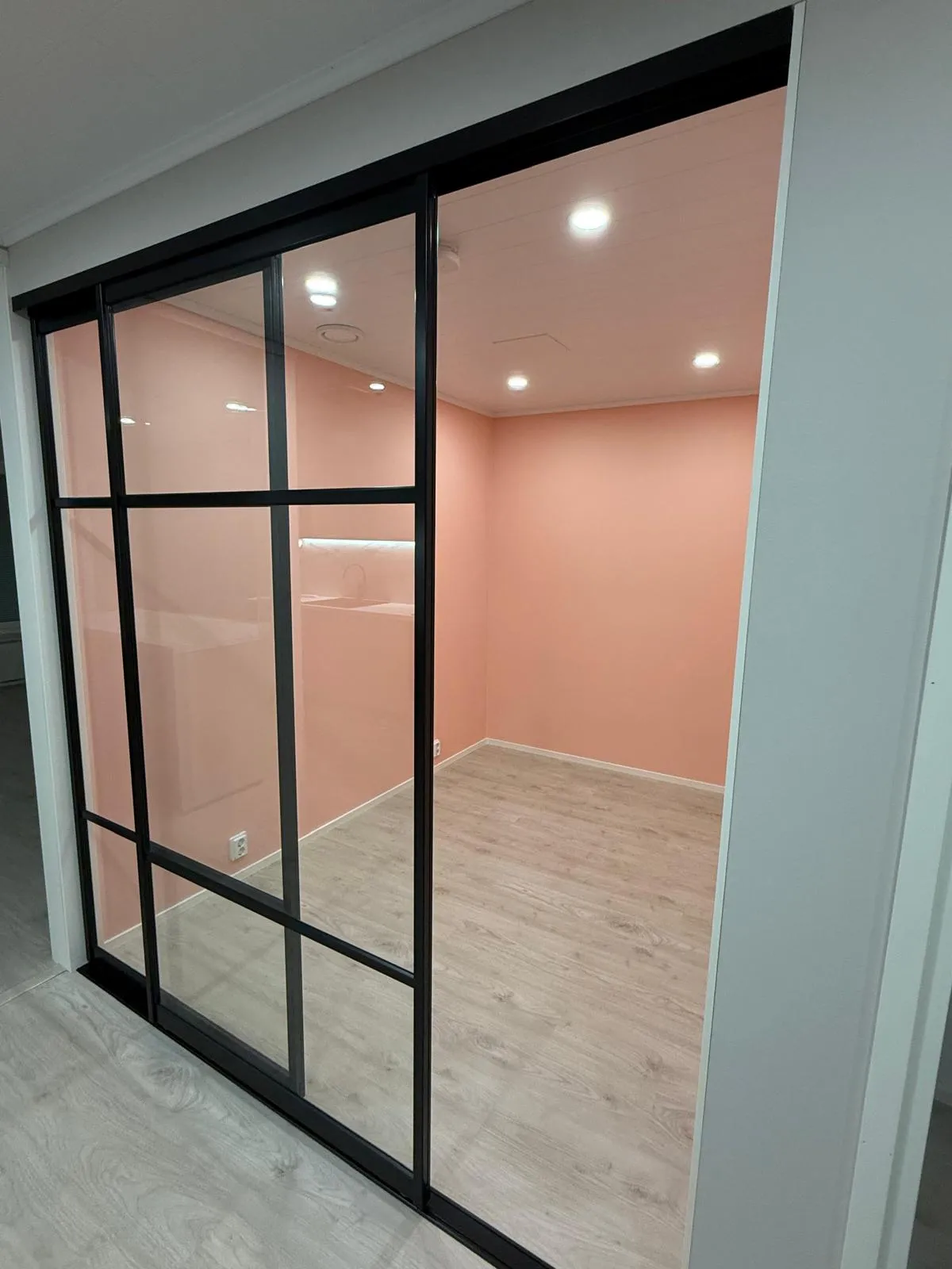 Kuva esittää modernia huonetta, jossa on mustakehyksinen lasinen liukuovijärjestelmä. Huoneen seinät ovat vaaleanpunaiset ja lattia vaaleaa puulaminaattia. Valkoisessa katossa on useita upotettuja valaisimia, ja takaseinällä on vaakasuora valonauha. Liukuovi on osittain auki, paljastaen huoneen sisätilan.