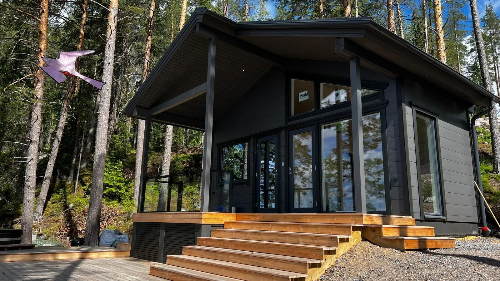 Moderni tummanharmaa mökki tai talo sijaitsee metsäisessä ympäristössä. Rakennuksessa on tumma harjakatto, suuret ikkunat, jotka heijastavat sinistä taivasta ja puita, sekä puinen terassi leveine portaineen. Ympärillä kasvaa tiheää mäntymetsää ja vihreää sammalta. Kuvan vasemmassa reunassa puussa roikkuu vaaleanpunainen leija. Oikealla näkyy kivikkoista maata ja hieman rakennustarvikkeita. Kuva on otettu kirkkaana, aurinkoisena päivänä.