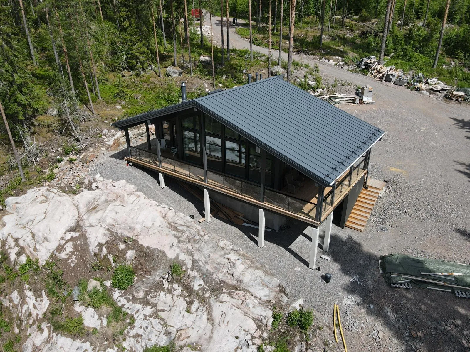 Ilmakuva modernista talosta metsäisessä, kallioisessa maastossa. Talossa on tummanharmaa peltikatto, suuri lasiseinä ja puinen terassi lasikaiteilla. Se on rakennettu rinteeseen ja sitä tukevat vaaleat betonipilarit. Terassilta laskeutuu puuportaat. Ympärillä on tiheää havumetsää, valkoisia kallioita ja soratie, jonka varrella on rakennustarvikkeita. Kuva on otettu kirkkaassa päivänvalossa, ja siinä näkyy selkeitä varjoja.