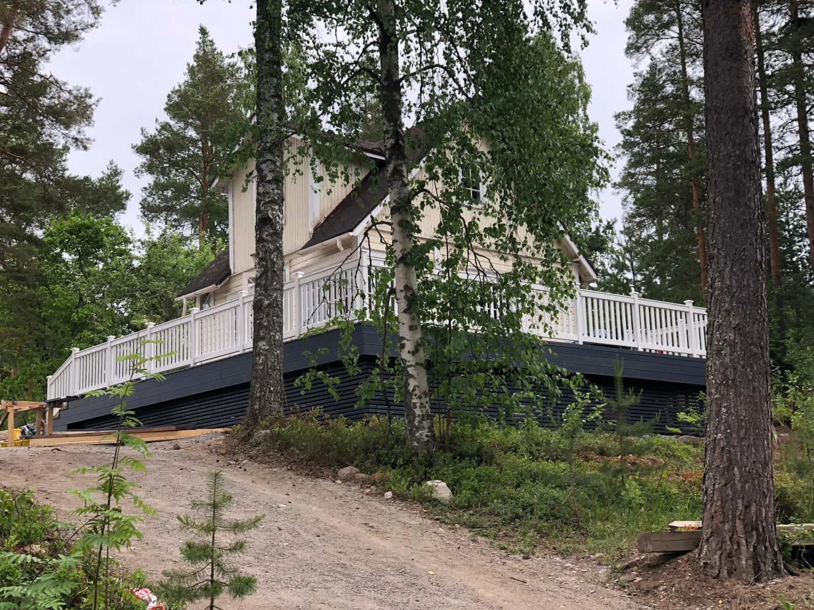 Vaaleankeltainen talo tummalla katolla ja valkoisella kaiteella varustetulla suurella terassilla sijaitsee metsäisessä rinteessä. Terassin tummanharmaa alaosa on näkyvissä. Edustalla on hiekkatie ja vihreää kasvillisuutta. Kuva on otettu pilvisenä päivänä, valaistus on tasainen.