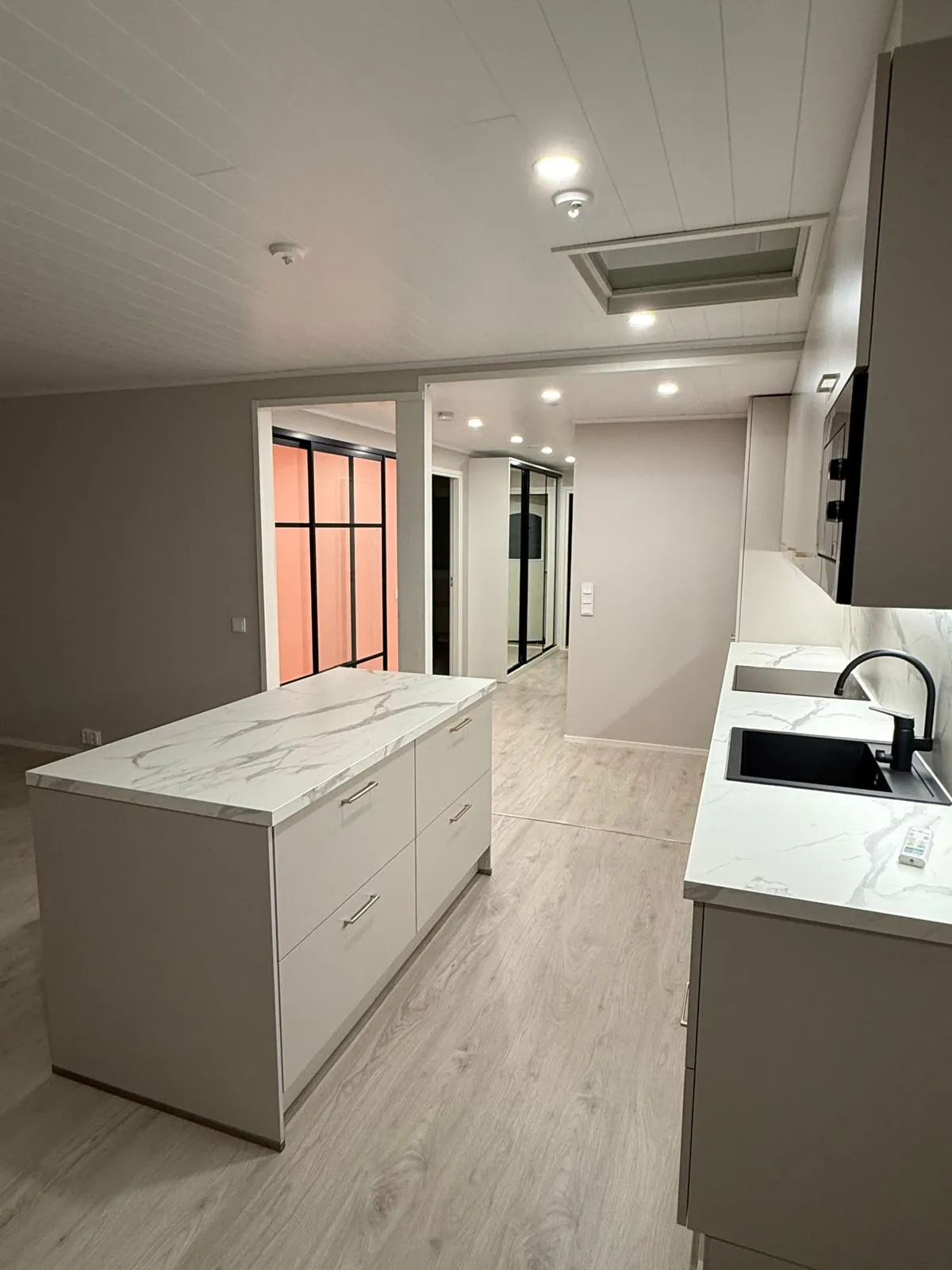 Vaaleanharmaa keittiö, jossa on valkoiset marmorikuvioiset työtasot ja vaalea puulattia. Edustalla on keittiösaareke neljällä laatikolla. Oikealla on musta allas ja hana sekä kaapin alusvalot. Katossa on valkoiset paneelit, upotettuja valoja, palovaroitin ja kattoikkuna. Taustalla näkyy käytävä, jossa on mustakehyksisiä liukuovia ja oranssinpunainen lasiseinä.