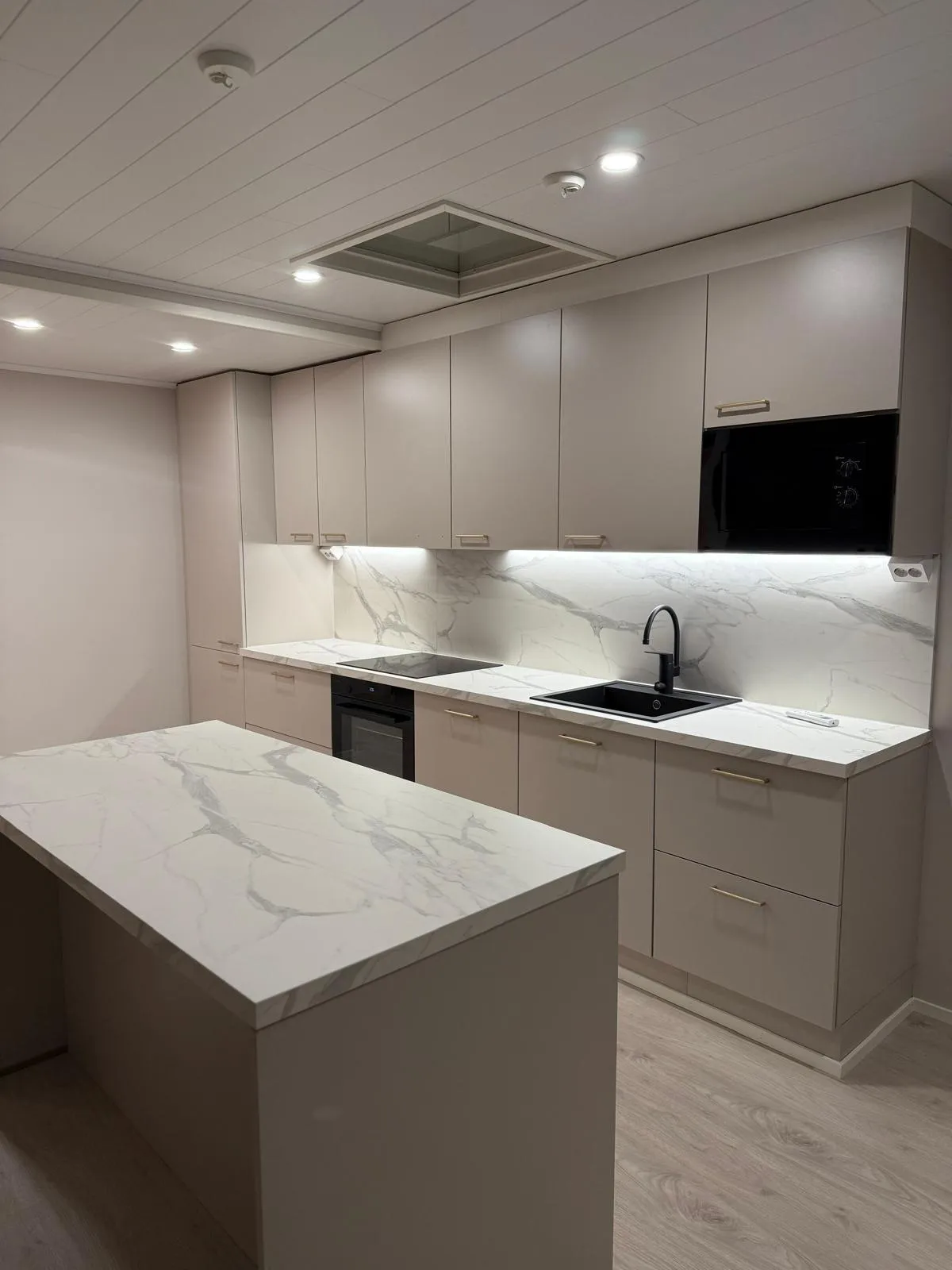 Moderni keittiö, jossa on vaaleanharmaat kaapit ja valkoiset marmorikuvioiset työtasot sekä välitila. Keittiössä on musta uuni, induktioliesi, pesuallas mustalla hanalla ja mikroaaltouuni. Kaappien alla on kirkas valaistus. Edustalla on samanlainen keittiösaareke. Katossa on valkoiset paneelit ja upotettuja valoja.
