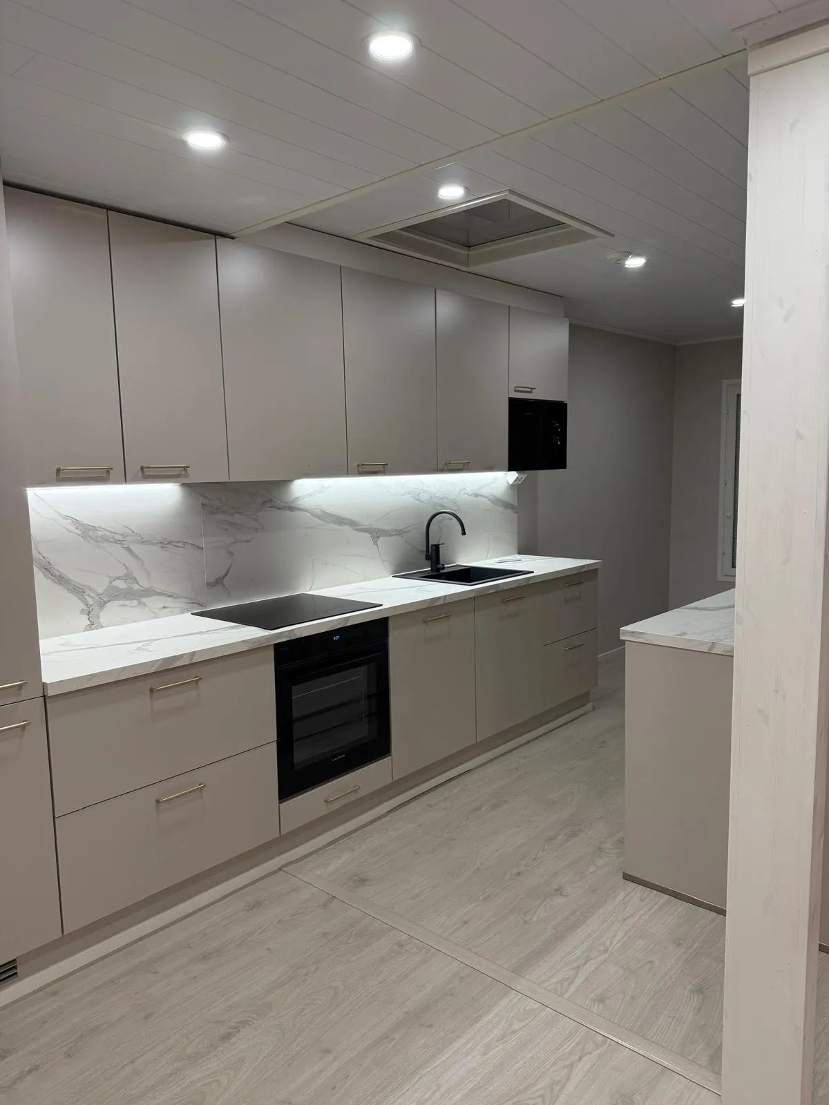 Moderni keittiö vaaleanbeigenvärisillä kaapeilla, valkoisella marmorikuvioisella työtasolla ja välitilalla. Musta liesitaso, uuni, mikroaaltouuni ja allas kultaisine vetimineen luovat kontrastia. Kaappien alla on kirkas valaistus, ja katossa on upotettuja valaisimia. Lattiana on vaalea puu.