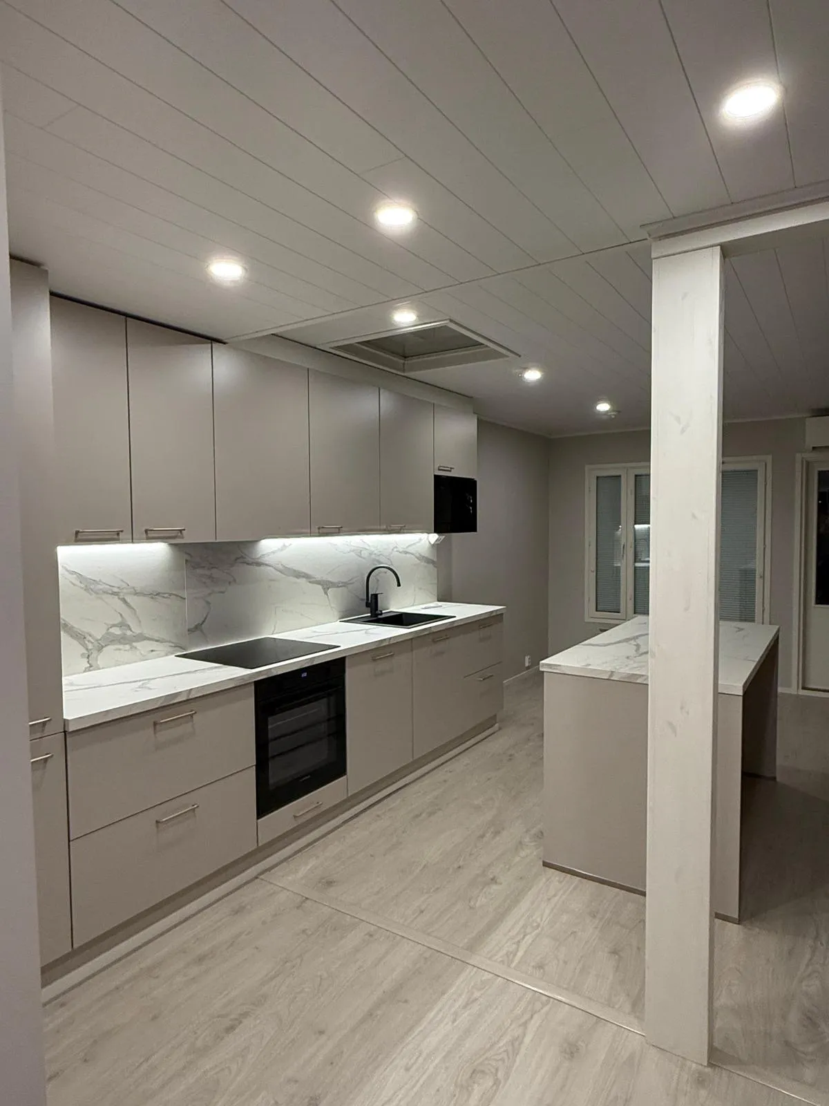 Moderni keittiö vaaleanharmailla kaapeilla ja marmorikuvioisilla valkoisilla työtasoilla. Musta liesi, uuni, tiskiallas, hana ja mikroaaltouuni. Katossa on upotettuja valoja ja kaappien alla kirkas valaistus. Vaalea puunvärinen laminaattilattia ja valkoinen paneelikatto, jossa on integroitu liesituuletin. Oikealla on valkoinen pylväs ja keittiösaareke.