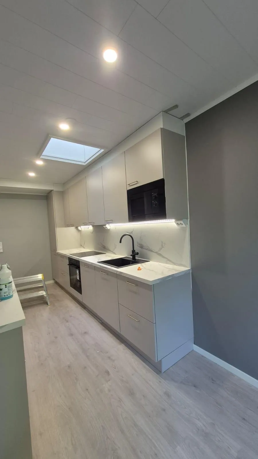 Moderni keittiö vaaleanharmailla kaapeilla, valkoisella marmorikuvioisella työtasolla ja välitilalla. Musta allas ja hana, sekä integroitu uuni ja mikroaaltouuni. Katossa on upotettuja valoja ja kattoikkuna, työtasoa valaisee led-nauha. Lattia on vaaleaa puunväristä laminaattia ja seinät harmaat. Vasemmalla näkyy tikkaat ja puhdistusainepullo.