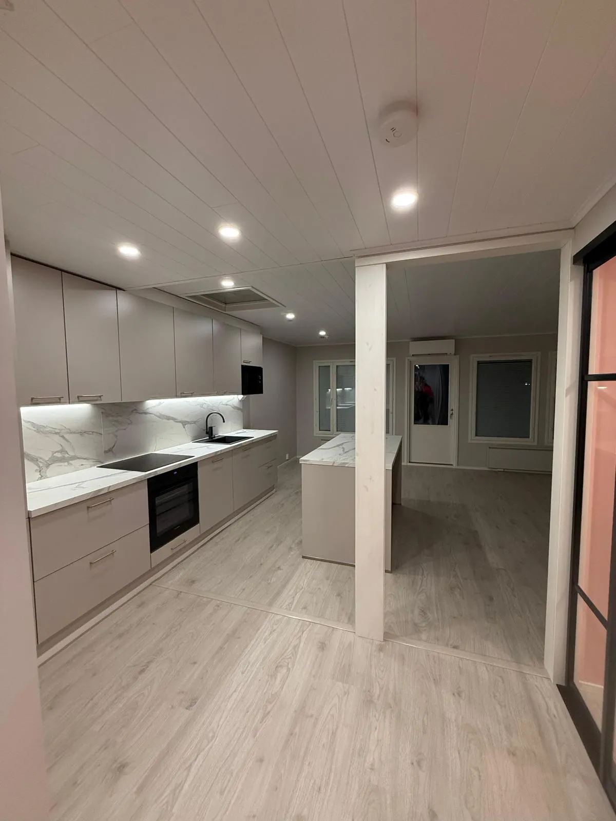 Moderni vaaleanharmaa keittiö, jossa on valkoiset marmorikuvioiset työtasot ja välitila, musta allas, liesi, uuni ja mikroaaltouuni. Keittiössä on upotetut katto- ja alakaappivalot. Vaalea puunvärinen laminaattilattia jatkuu viereiseen tyhjään huoneeseen, jossa on ikkunat ja ovi. Valkoisessa paneelikatossa on palovaroitin ja upotettuja valoja.