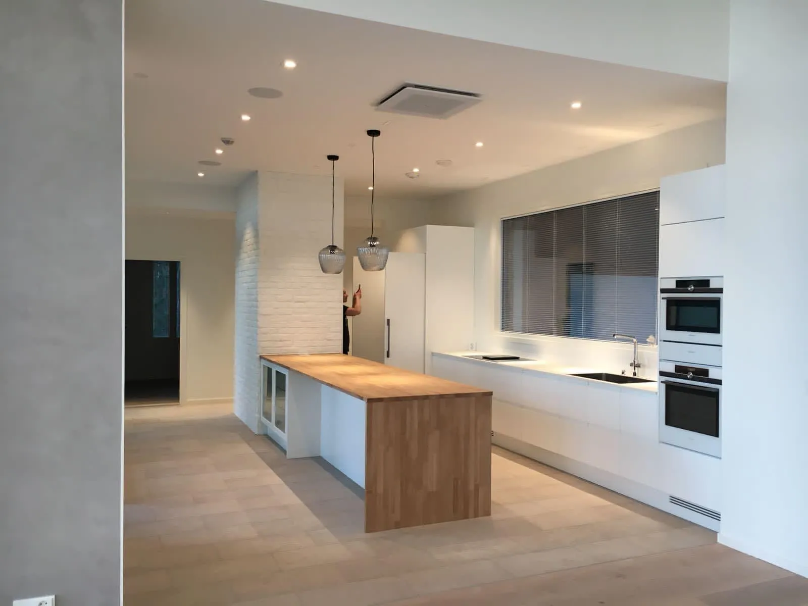 Moderni keittiö, jossa on valkoiset kaapit ja vaalea puulattia. Keskellä on pitkä keittiösaareke, jossa on puinen työtaso ja valkoinen alaosa. Saarekkeen yläpuolella roikkuu kaksi lasista riippuvalaisinta. Katossa on useita upotettuja valoja ja ilmastointiyksikkö. Takaseinällä on suuri ikkuna tummanharmailla sälekaihtimilla, musta allas ja kromihana. Oikealla on pystysuoraan pinotut integroidut uuni ja mikroaaltouuni. Vasemmalla on valkoinen jääkaappi. Yksi seinä on valkoista tiiltä. Henkilö näkyy osittain saarekkeen takana. Huone on kirkkaasti valaistu.