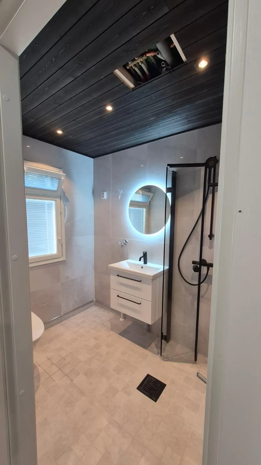 Moderni kylpyhuone, jossa on tumma puupaneelikatto upotetuilla valoilla ja avoin huoltoluukku putkineen. Seinät ovat vaaleanharmaata laattaa ja lattia vaaleanbeigeä neliölaattaa mustalla lattiakaivolla. Huoneessa on ikkuna sälekaihtimilla, valkoinen allaskaappi mustalla hanalla ja pyöreä taustavalaistu peili. Oikealla näkyy suihkutila mustakehyksisellä lasiseinällä ja mustalla suihkulla.