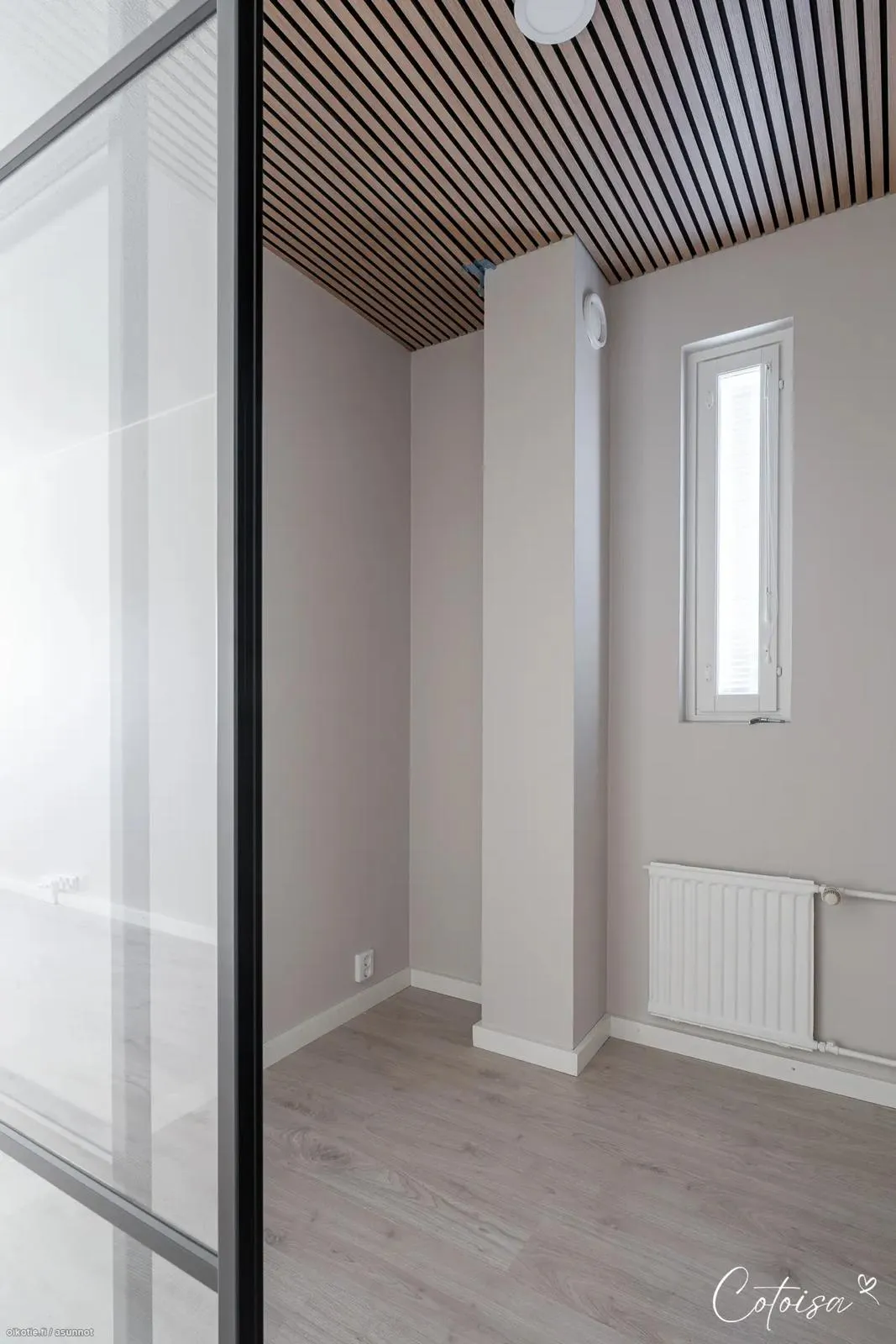 Tyhjä huone, jossa on moderni sisustus. Vasemmalla näkyy mustakehyksinen lasiseinä. Katossa on vaalea puurimakangas mustilla raidoilla ja pyöreä valkoinen kattovalaisin. Seinät ja keskellä oleva pylväs ovat vaalean harmaanbeigen sävyiset. Pylväässä on valkoinen ilmanvaihtoventtiili. Oikealla seinällä on kapea valkoinen ikkuna ja sen alapuolella valkoinen patteri. Lattialla on vaalea puunvärinen laminaatti ja valkoiset jalkalistat. Huoneessa on kirkas valaistus.