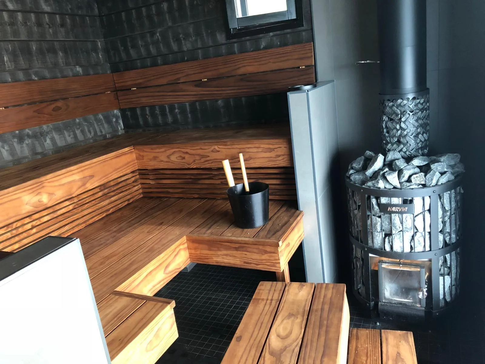 Moderni sauna, jossa on tummat puuseinät ja vaaleanruskeat, L-muotoiset puulauteet. Oikealla on suuri, musta Harvia-kiuas täynnä kiviä ja musta savupiippu. Lauteilla on musta ämpäri ja kaksi puista löylykauhaa. Lattialla on tummat laatat. Ylhäällä seinässä näkyy pieni ikkuna. Tila on kirkkaasti valaistu.