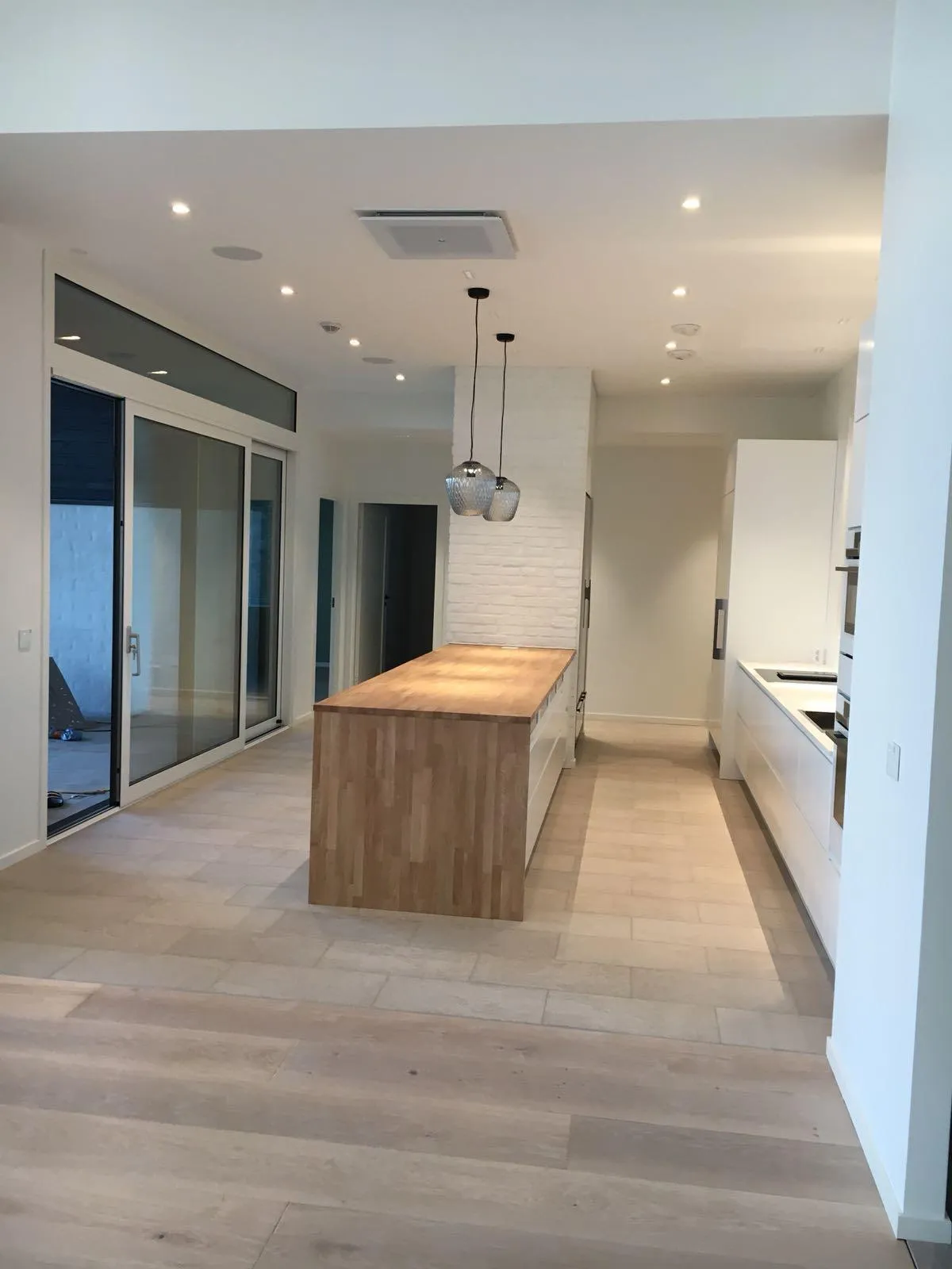 Valoisa ja moderni keittiö, jossa on vaalea puulattia ja laattalattia. Keskellä on suuri keittiösaareke vaalealla puutasolla ja valkoisilla kaapeilla. Saarekkeen yläpuolella roikkuu kaksi lasista riippuvalaisinta. Oikealla on valkoinen keittiölinjasto integroiduilla laitteilla ja pesualtaalla. Vasemmalla on suuret lasiset liukuovet. Katossa on upotettuja spotteja ja ilmastointilaite. Saarekkeen takana on valkoinen tiiliseinä.