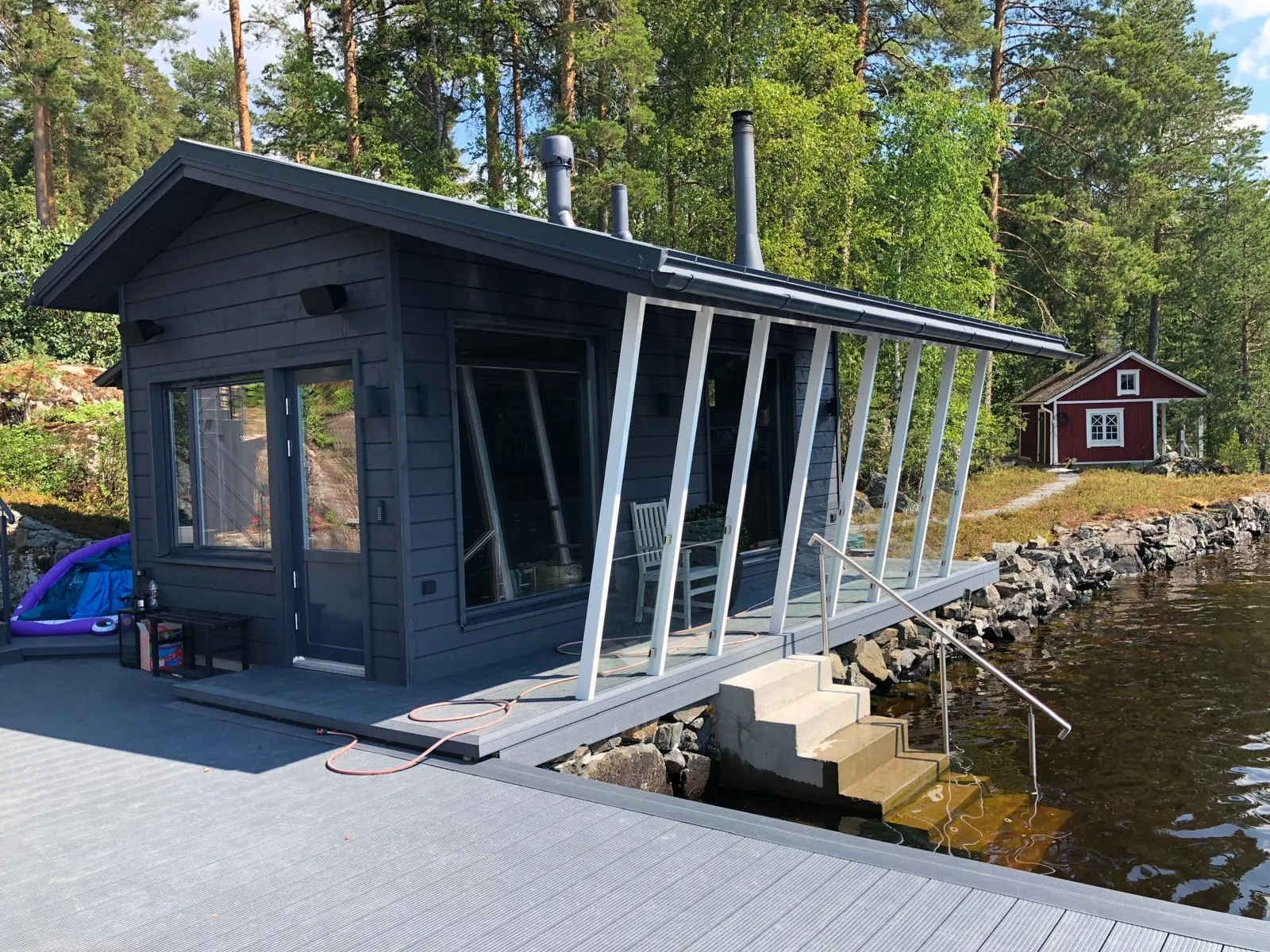 Aurinkoisena päivänä tummanharmaa mökki, jossa on suuret ikkunat ja valkoiset vinot tukipylväät, seisoo harmaan laiturin äärellä. Laiturilla on oranssi letku ja pieni penkki. Betonirappuset metallikaiteineen laskeutuvat tummaan veteen. Taustalla puiden ympäröimänä on pieni punainen mökki. Mökissä on kolme tummaa savupiippua.