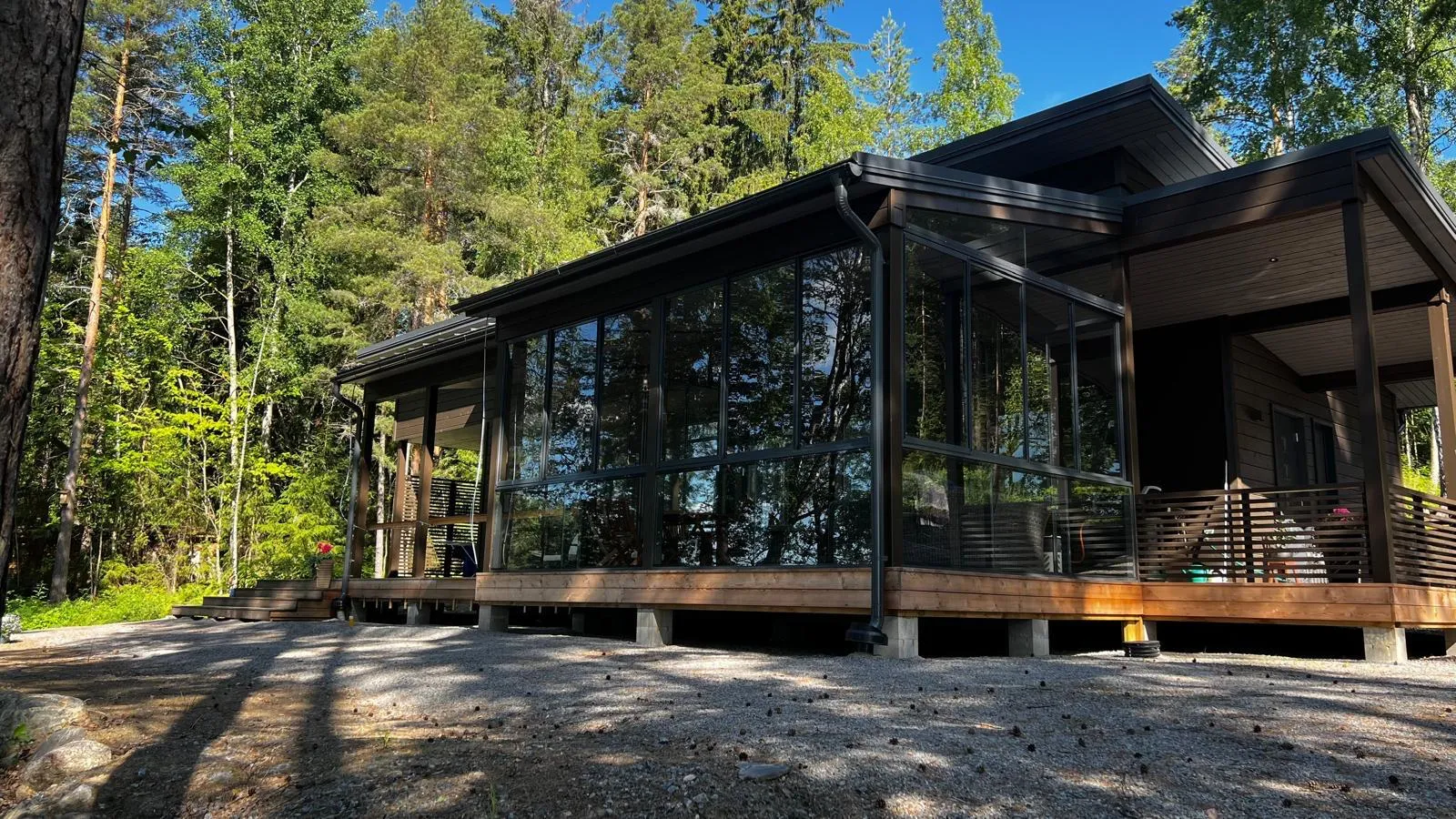 Moderni tummanruskea mökki metsän keskellä. Mökissä on suuri lasitettu terassi, joka heijastaa vihreitä puita ja kirkasta sinistä taivasta. Puinen terassi ja portaat johtavat mökkiin, joka on korotettu betonipilareiden varaan. Edustalla on sorapiha, jossa on käpyjä ja auringon luomia varjoja.