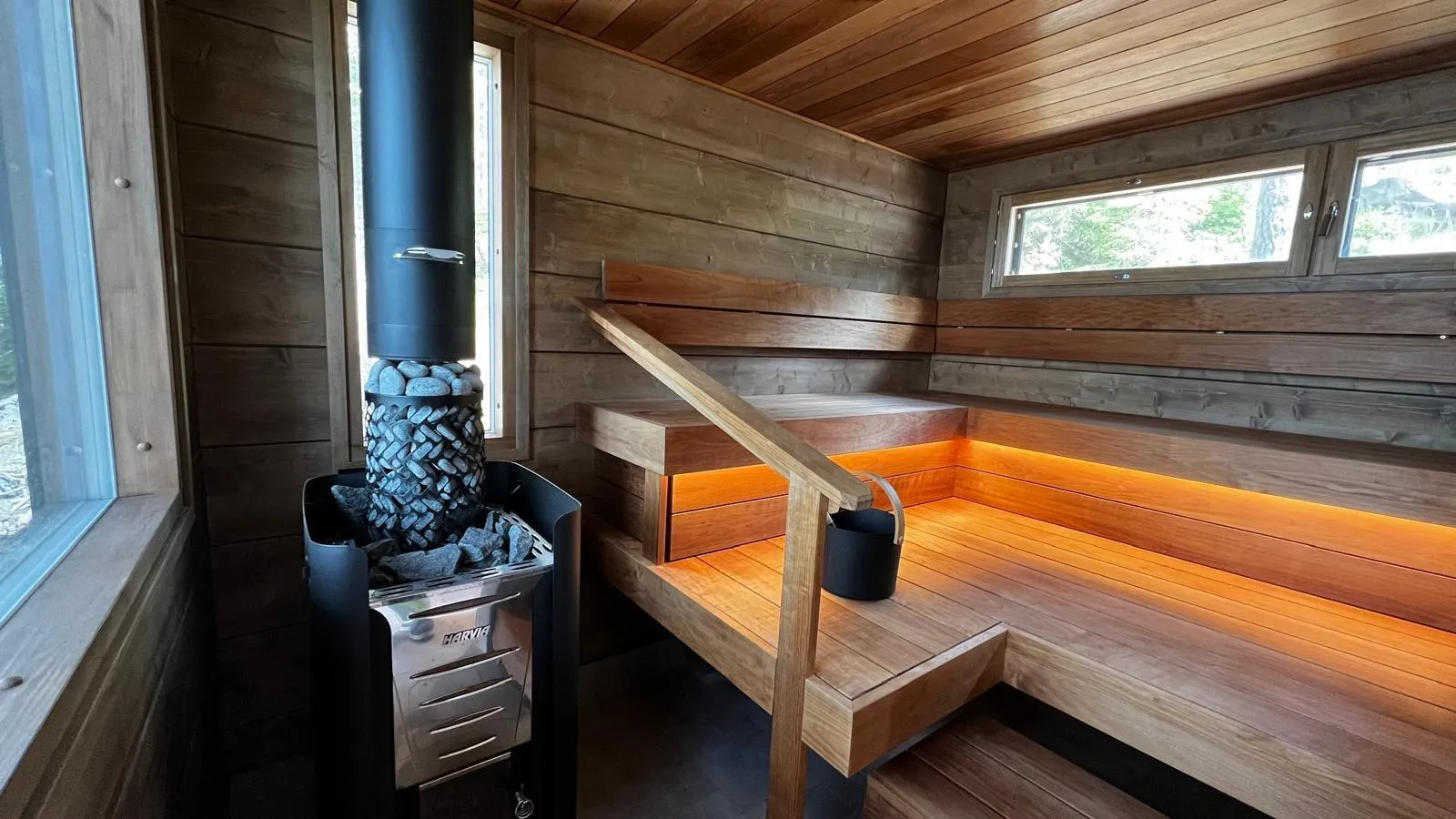 Moderni puusauna, jossa on tummanruskeat puuseinät ja -katto. Vasemmalla on musta Harvia-kiuas kiuaskivineen ja korkea savupiippu. Oikealla on puiset lauteet, joissa on lämmin oranssi epäsuora valaistus. Lauteille johtaa puinen käsinoja, josta roikkuu musta ämpäri. Ikkunoista näkyy metsää.