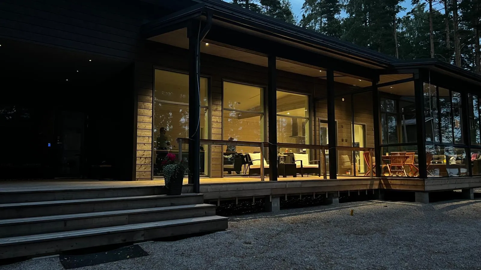 Moderni tumma puutalo iltahämärässä, jossa on suuria ikkunoita ja valaistu puinen terassi portaineen. Ikkunoista heijastuu sisätilojen lämmin keltainen valo, joka valaisee huonekaluja ja luo kutsuvan tunnelman. Oikealla on lasitettu tila. Taustalla näkyy tumma metsä ja hämärtyvä taivas.