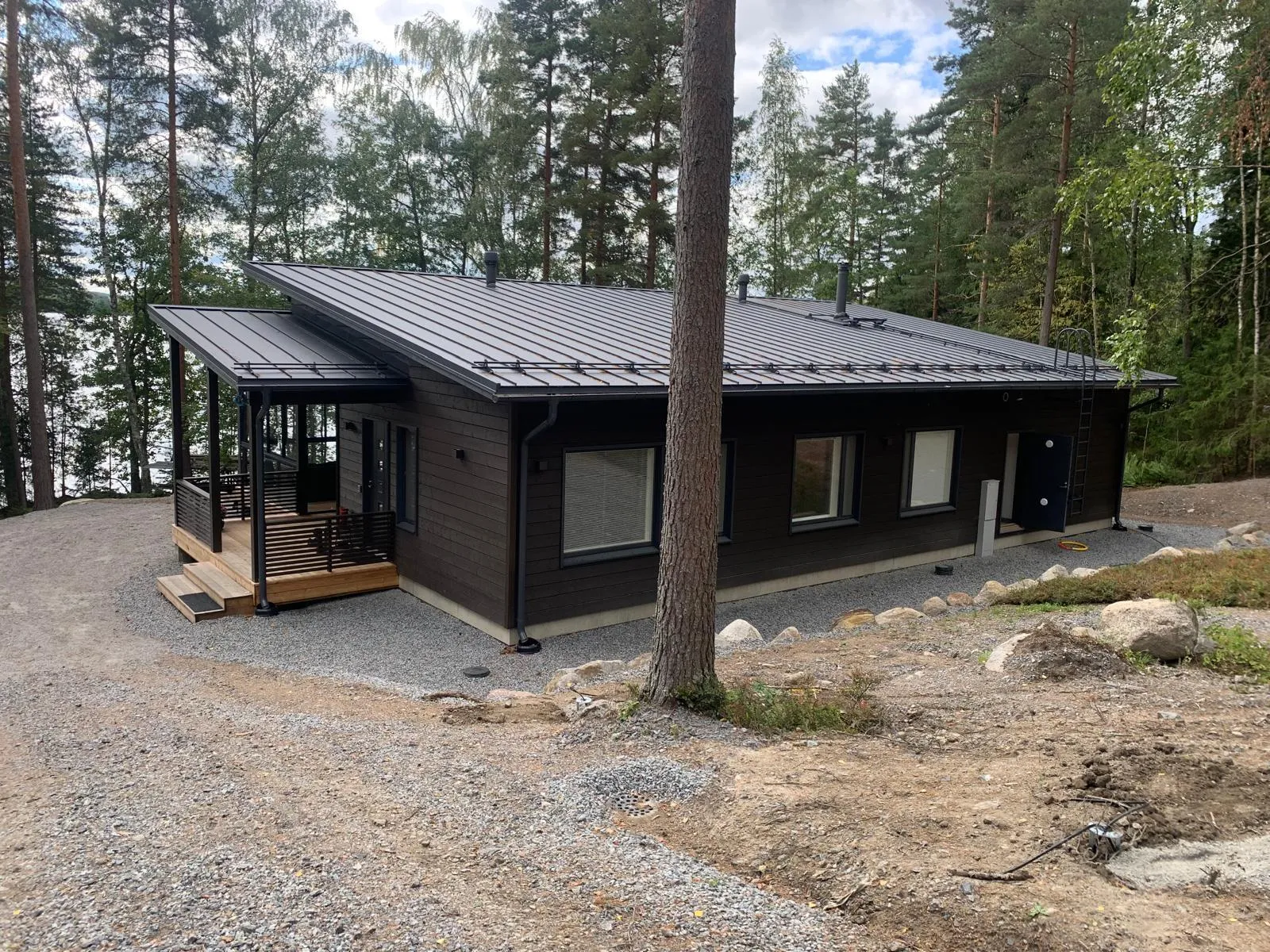 Tummanruskea puutalo tummanharmaalla peltikatolla ja mustilla räystäskouruilla metsäisessä maisemassa. Vasemmalla on katettu puukuisti mustilla kaiteilla. Oikealla puolella on tumma ovi ja seinätikkaat. Talon ympärillä on soraa ja maata, ja taustalla näkyy järvi. Korkeat puut ympäröivät rakennusta. Pilvinen päivä valaisee kuvaa.