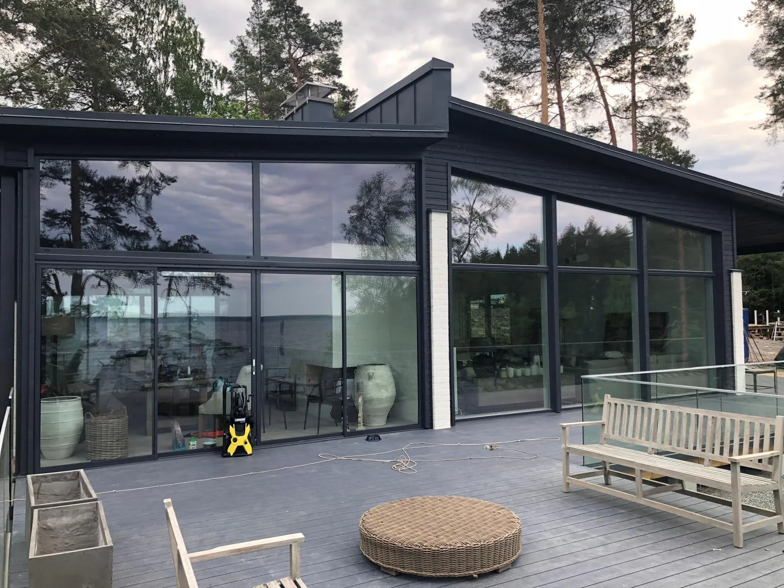 Moderni tumma talo suurilla ikkunoilla, jotka heijastavat pilvistä taivasta ja puita. Edustalla on harmaa puuterassi, jossa on puinen penkki, rottinkinen pöytä, puutuoli, keltainen painepesuri ja betoniruukkuja. Ikkunoiden läpi näkyy vesistö.