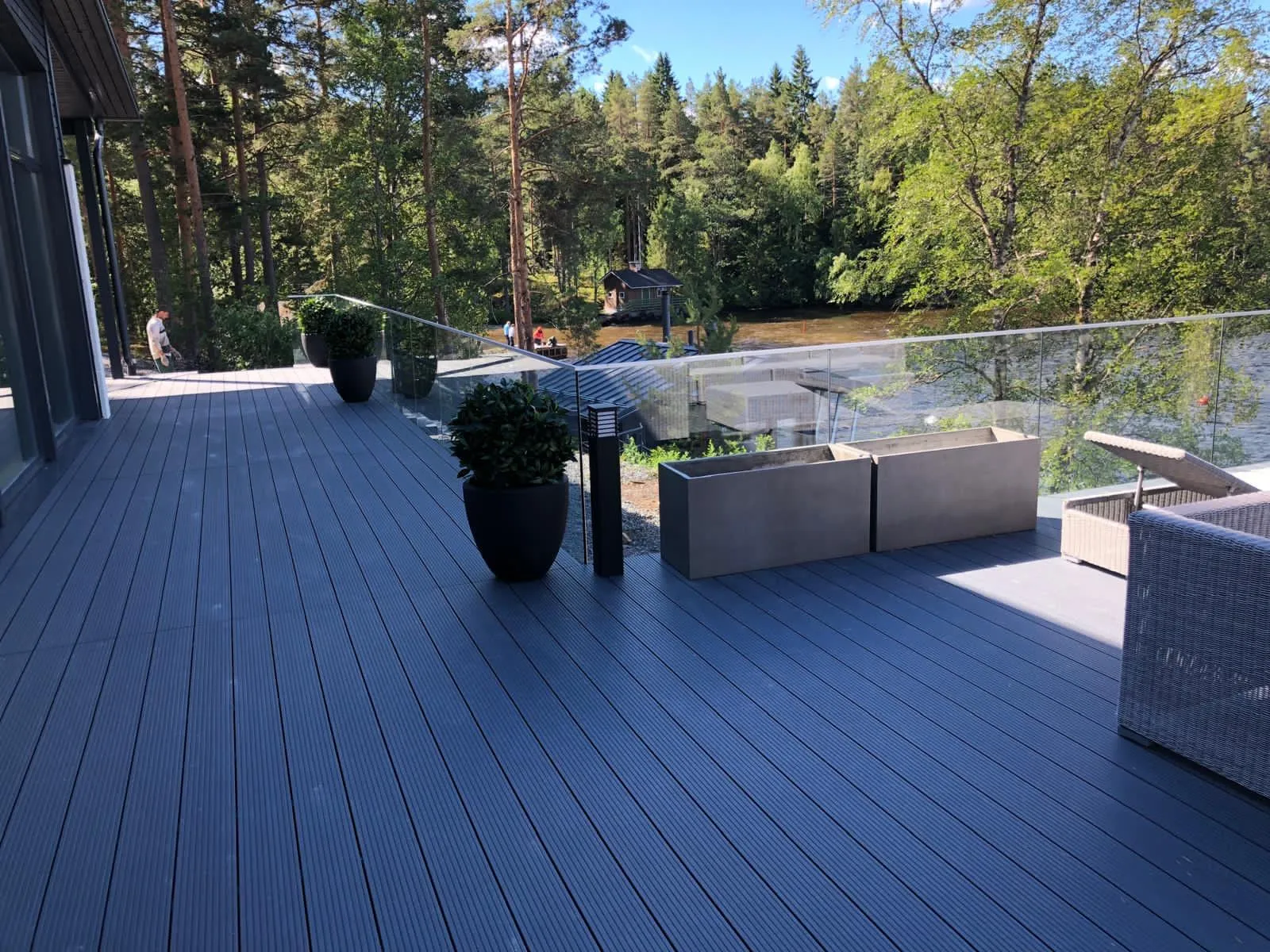 Aurinkoinen kuva tummanharmaasta terassista, jossa on lasikaide ja näkymä joelle tai järvelle. Terassilla on tummia ruukkuja vihreine pensaineen, kaksi suorakaiteen muotoista vaaleanharmaata betonista istutusastiaa, musta ulkovalo ja rottinkikalusteita. Veden toisella puolella on tiheä metsä, pieni puumökki ja ihmisiä. Kirkas auringonvalo luo varjoja terassille.
