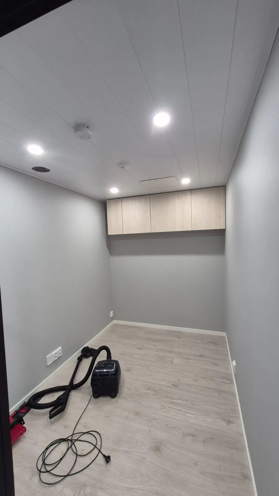 Tyhjä huone, jossa on vaaleanharmaat seinät ja vaalea puunvärinen laminaattilattia. Valkoisessa paneelikatossa on useita kirkkaita upotettuja valoja, palovaroitin ja ilmanvaihtoritilä. Yhdellä seinällä on rivi vaaleita puunvärisiä yläkaappeja. Lattialla on musta imuri letkuineen ja virtajohdoineen, ja vasemmalla näkyy punainen harja.