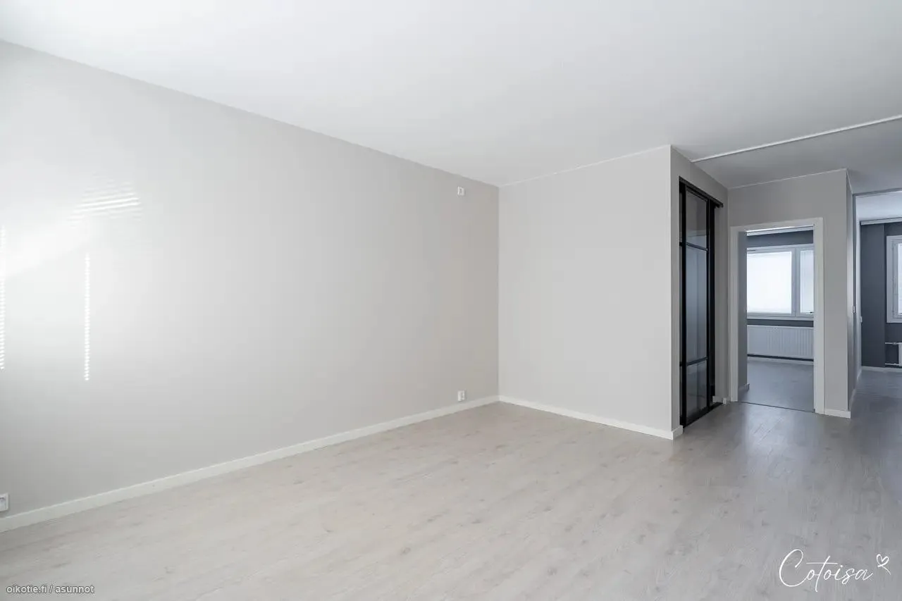 Tyhjä, vaaleanharmaa huone, jossa on vaalea puulattia. Vasemmalla seinällä on ikkuna, josta tulee raitainen valo sisään. Oikealla on mustakehyksinen lasiovi ja avoin oviaukko toiseen huoneeseen, jossa näkyy ikkuna ja patteri.