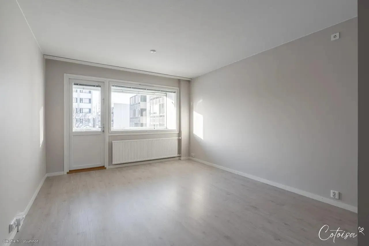 Tyhjä, valoisa huone vaaleanharmailla seinillä ja vaalealla puulattialla. Huoneessa on valkoinen ovi ja suuri ikkuna, jonka alla on valkoinen patteri. Ikkunasta näkyy kaupunkimaisemaa. Luonnonvalo luo heijastuksia lattialle ja kirkkaan kohdan oikealle seinälle.