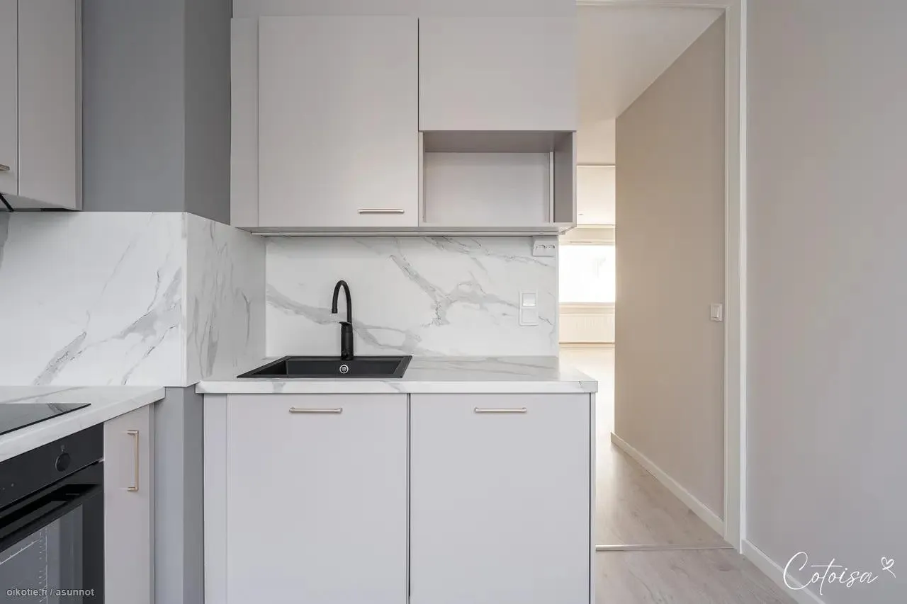 Moderni vaaleanharmaa keittiö, jossa on valkoinen marmorikuvioinen välitila ja työtaso. Musta allas ja hana ovat keskellä. Ylä- ja alakaapit ovat vaaleanharmaat hopeisilla vetimillä. Vasemmalla näkyy osa uunista. Oikealla avautuu vaalea käytävä, josta näkyy ikkuna ja valoisa huone. Lattia on vaaleaa puuta.