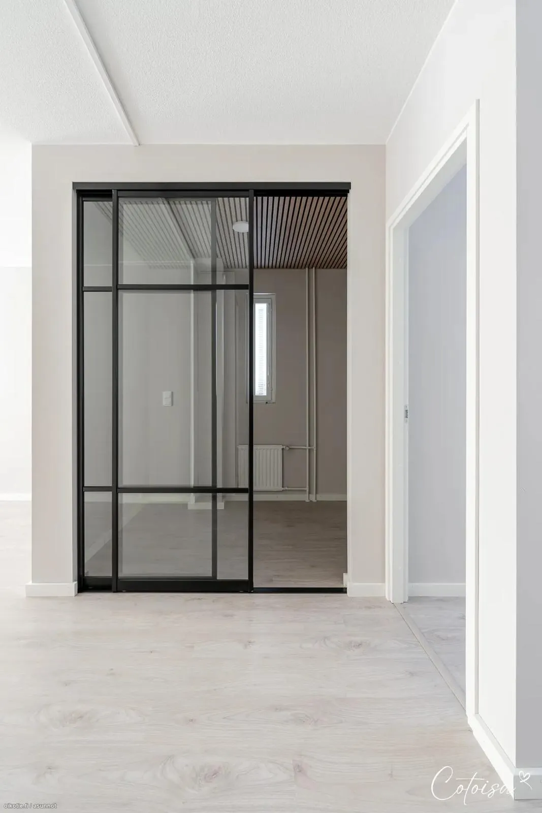 Valoisa, tyhjä sisätila, jossa on vaalea puulattia ja valkoiset seinät. Keskellä on mustakehyksinen lasiliukuovi, joka on osittain auki ja paljastaa huoneen, jossa on vaaleanharmaat seinät, puurimakatto, kapea ikkuna ja patteri. Oikealla on valkoinen ovenkarmi.