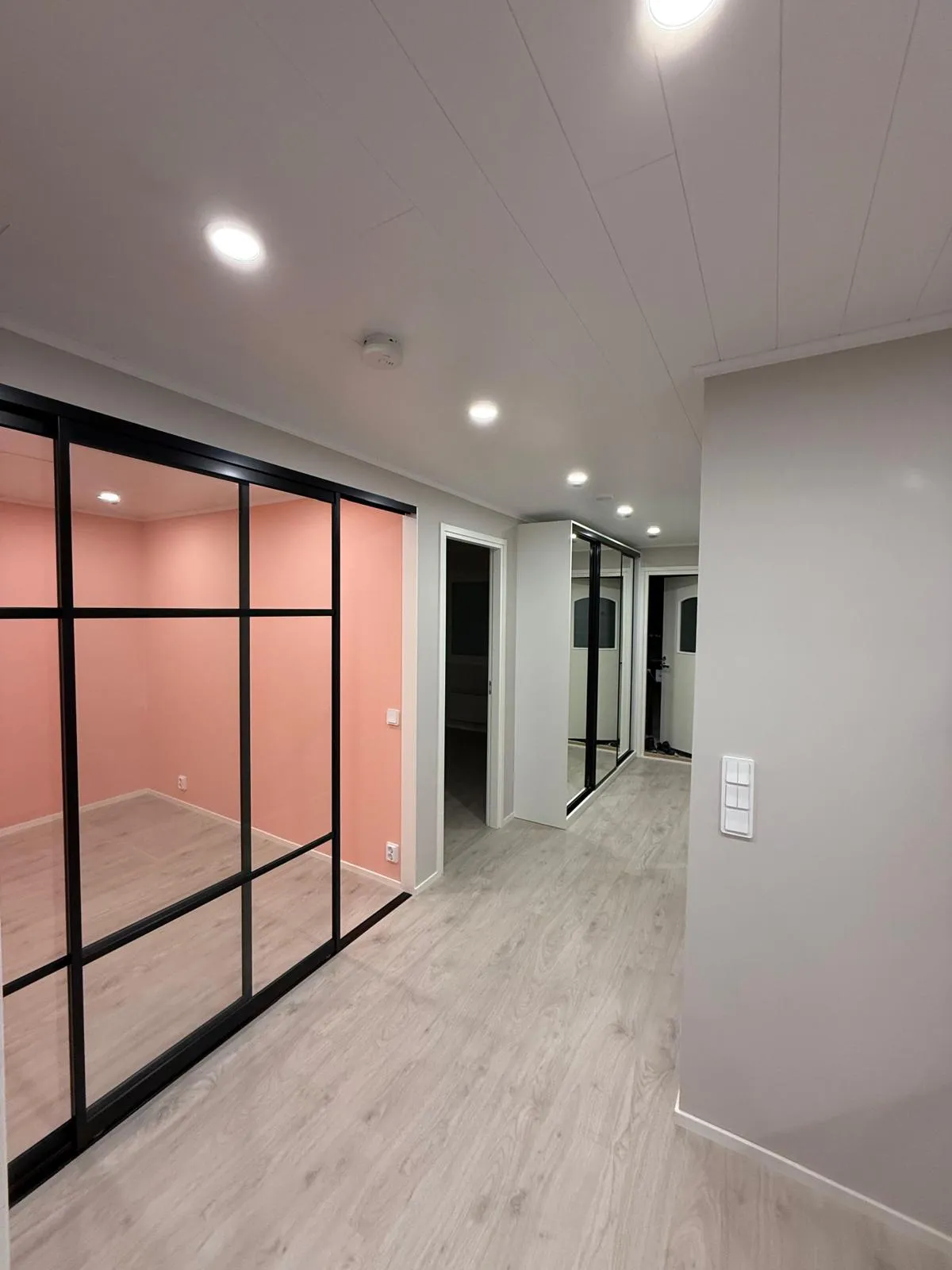 Käytävä, jossa vaaleat puulattiat ja vaaleanharmaat seinät. Vasemmalla on huone vaaleanpunaisilla seinillä ja mustakehyksisillä lasiliukuovilla. Katossa on valkoiset paneelit, useita upotettuja spotvaloja ja palovaroitin. Oikealla seinällä on valkoinen kolmoisvalokatkaisin. Käytävällä edempänä näkyy avoimia oviaukkoja ja valkoinen peililiukuovikaappi. Tila on kirkkaasti valaistu.