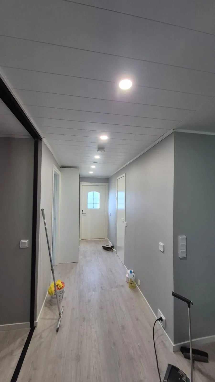 Pitkä, vaaleanharmaa käytävä, jossa on valkoinen paneelikatto ja vaalea puunvärinen lattia. Katossa on useita päällä olevia upotettuja kohdevaloja ja palovaroitin. Käytävän päässä on valkoinen ovi ruudullisella ikkunalla. Vasemmalla seinällä on valokatkaisija ja lattialla moppi sekä ämpäri keltaisine käsineineen. Oikealla seinällä on pistorasia ja valokatkaisija, ja lattialla näkyy puhdistusainepulloja ja musta sähköjohto.